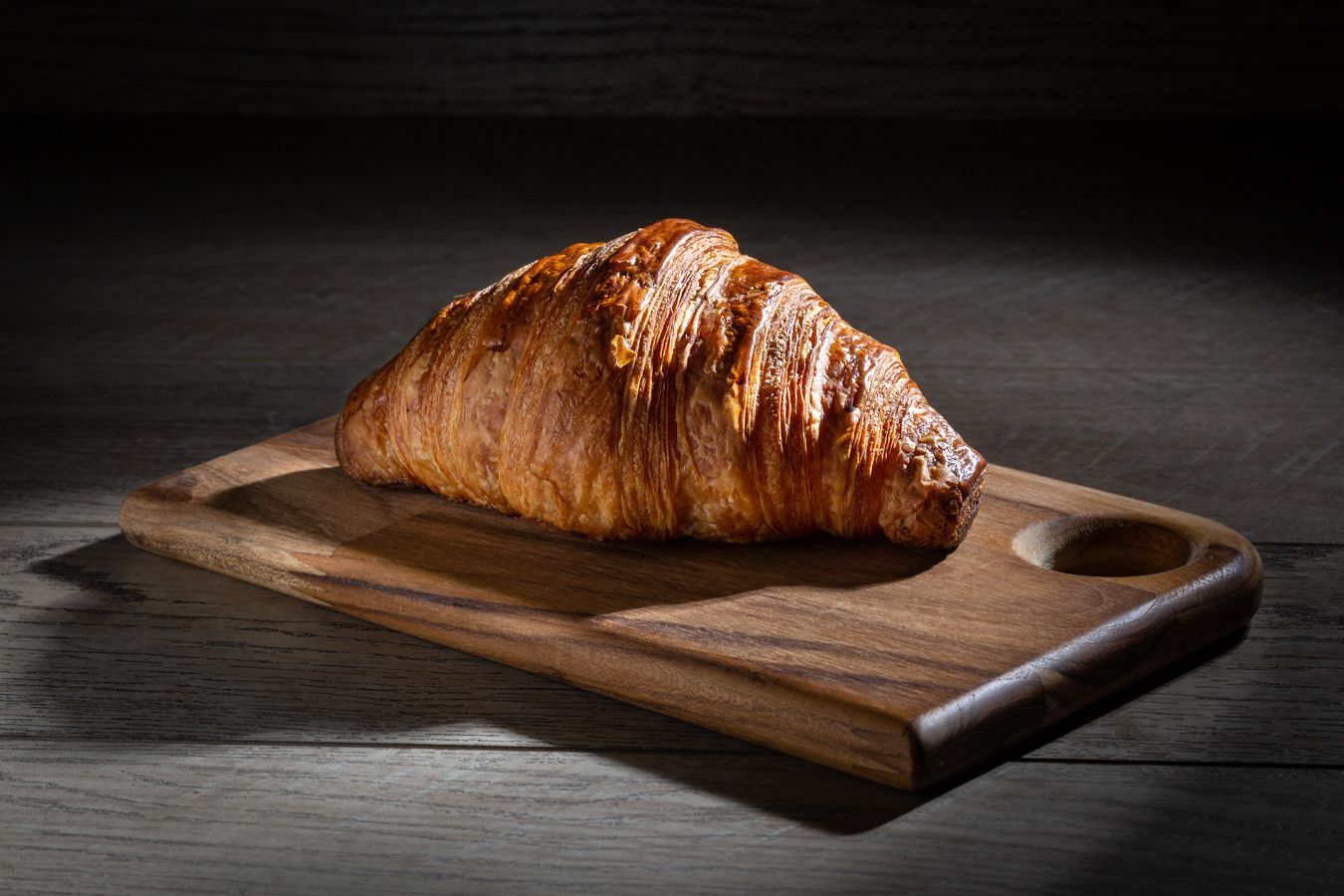 Croissant dourado sobre uma tábua de madeira contra um fundo escuro.