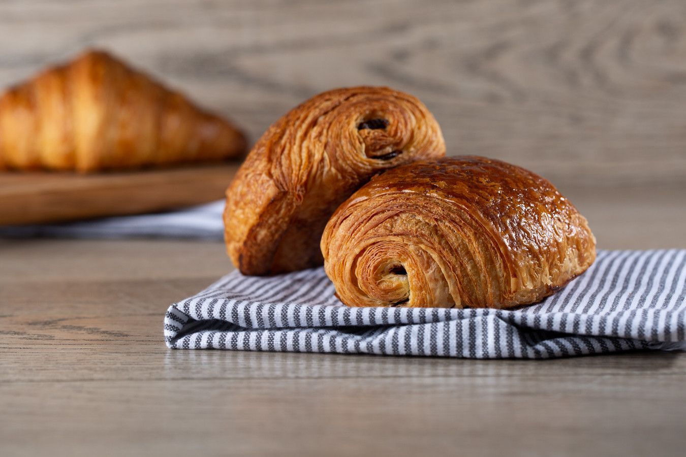 Dois croissants sobre uma toalha xadrez, com um terceiro croissant desfocado ao fundo, sobre uma mesa.