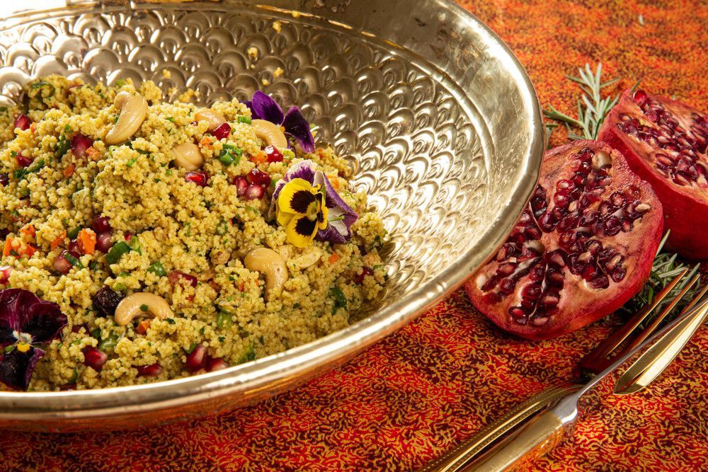 Tigela decorativa de cuscuz com romã, flores e especiarias sobre uma toalha de mesa vermelha.