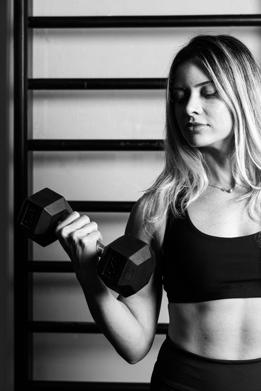 Mulher segurando um haltere em uma academia, em pé ao lado de barras na parede, usando um top esportivo preto.