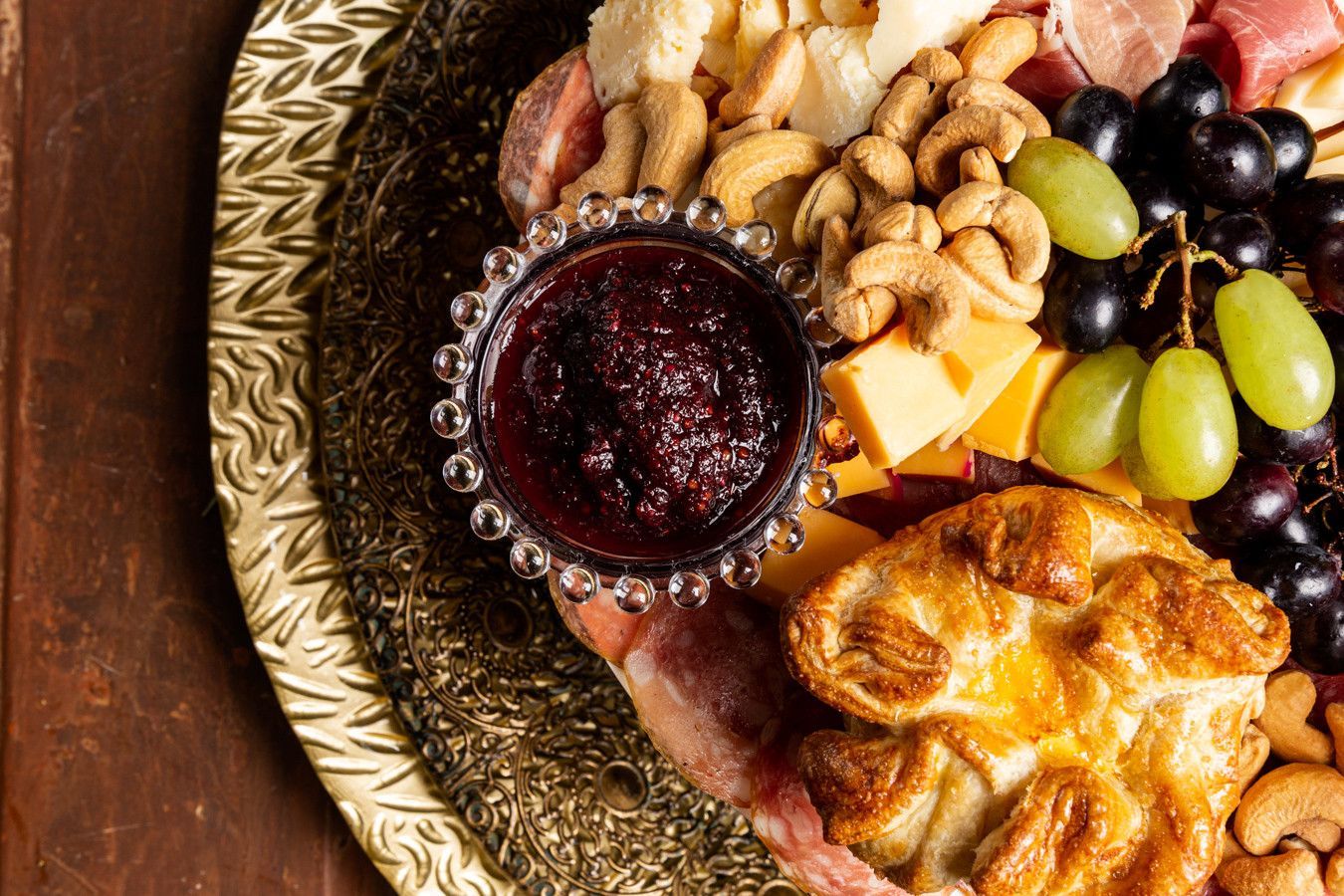 Tábua de charcutaria com geleia, uvas, queijo, biscoitos e nozes em um prato decorativo dourado.