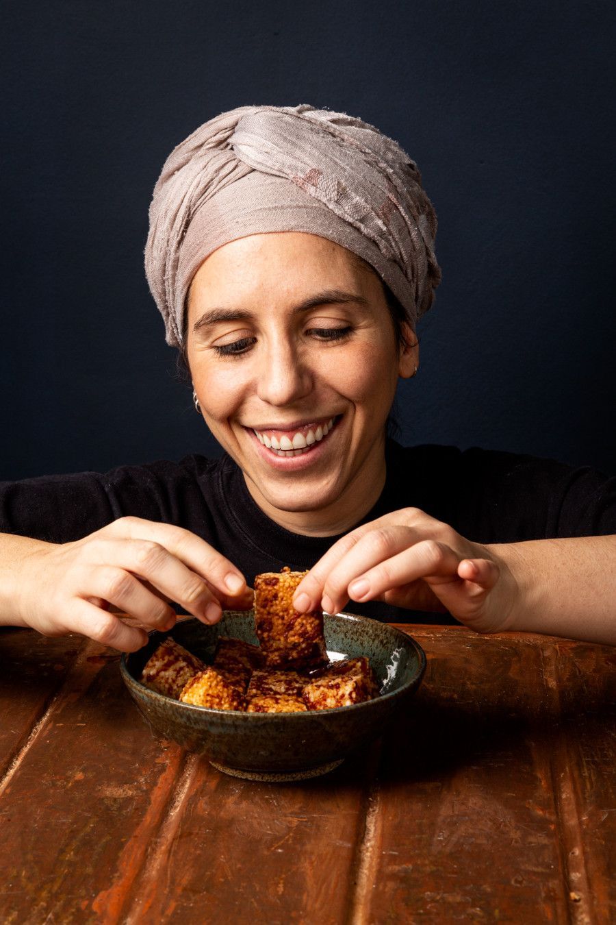 Pessoa sorridente com um turbante de toalha comendo em uma mesa de madeira.