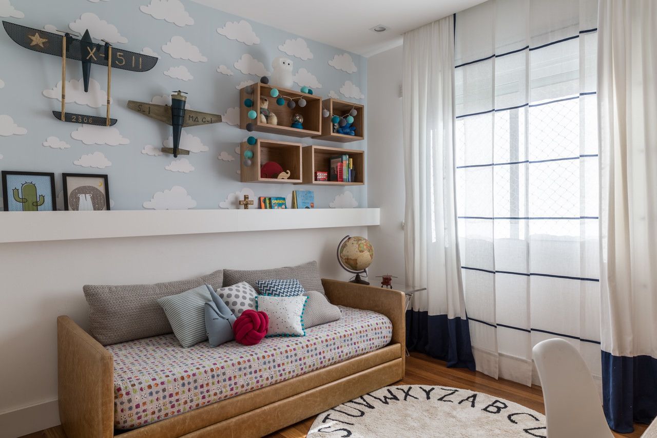Quarto infantil luminoso com uma cama de solteiro bege, prateleiras na parede e uma grande janela com cortinas brancas.