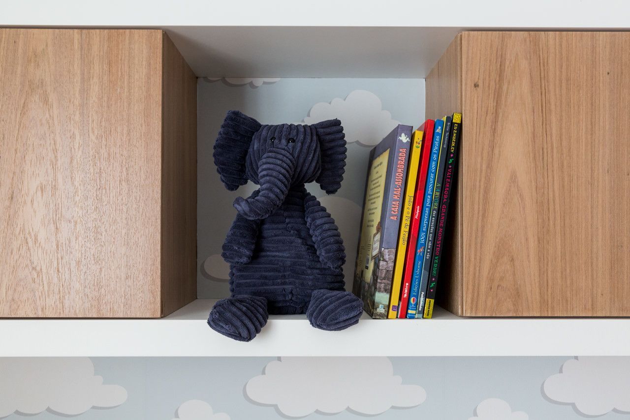 Elefante de pelúcia azul-escuro em uma prateleira ao lado de livros coloridos, entre portas de armário de madeira.