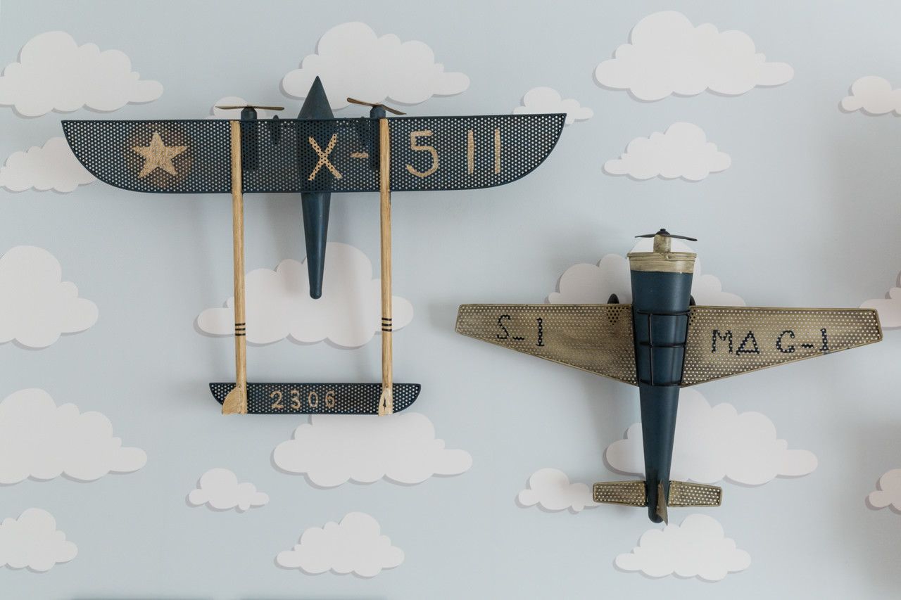 Dois modelos de aviões antigos montados em uma parede com estampa de nuvens, um azul escuro e outro bege com uma figura de piloto.