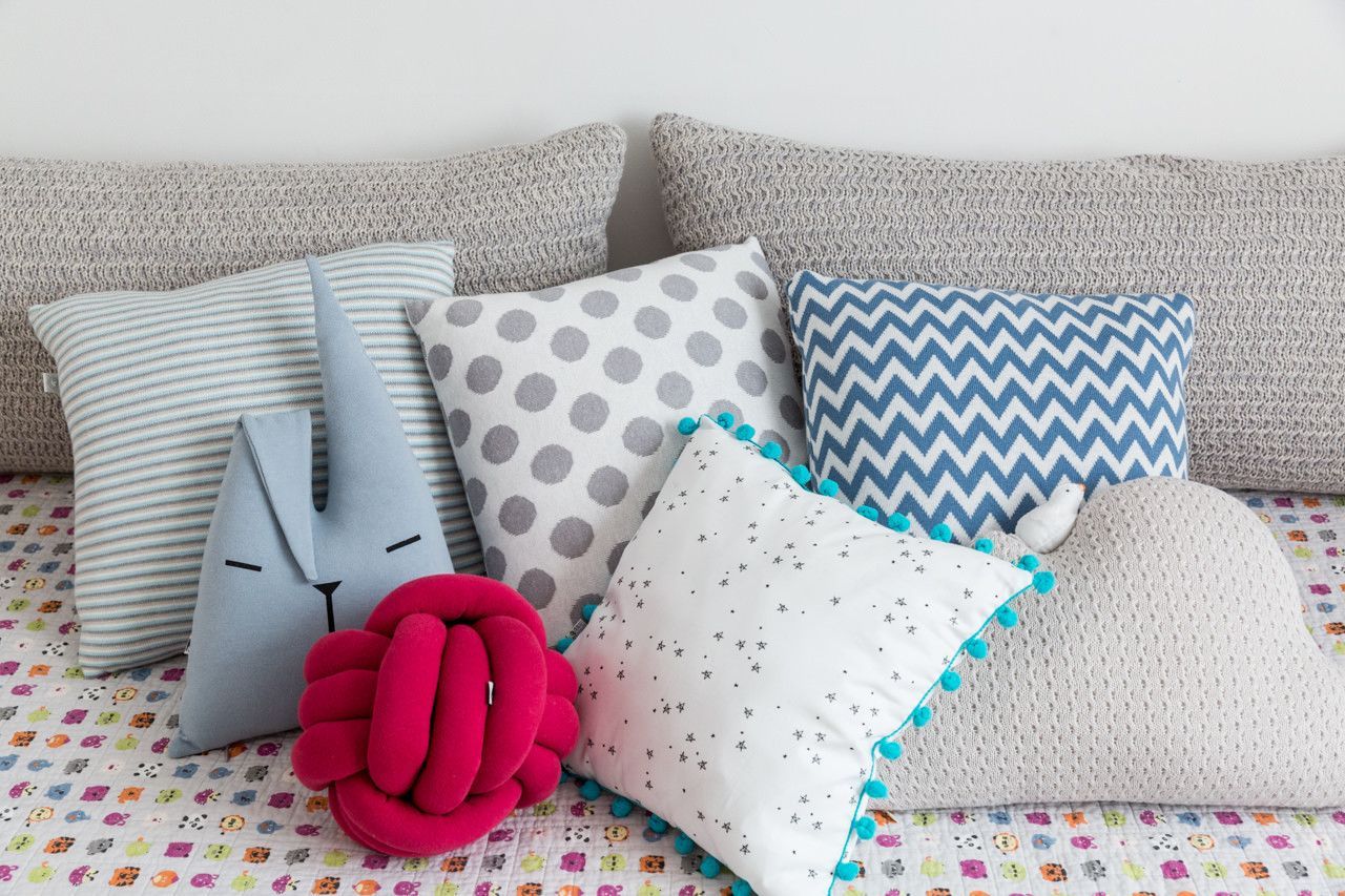 Almofadas estampadas e uma almofada rosa com nó dispostas sobre uma cama encostada em uma cabeceira estofada cinza.