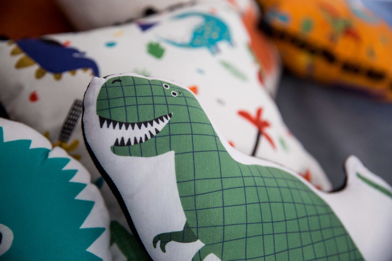 Dinossauro de pelúcia verde entre almofadas coloridas estampadas.