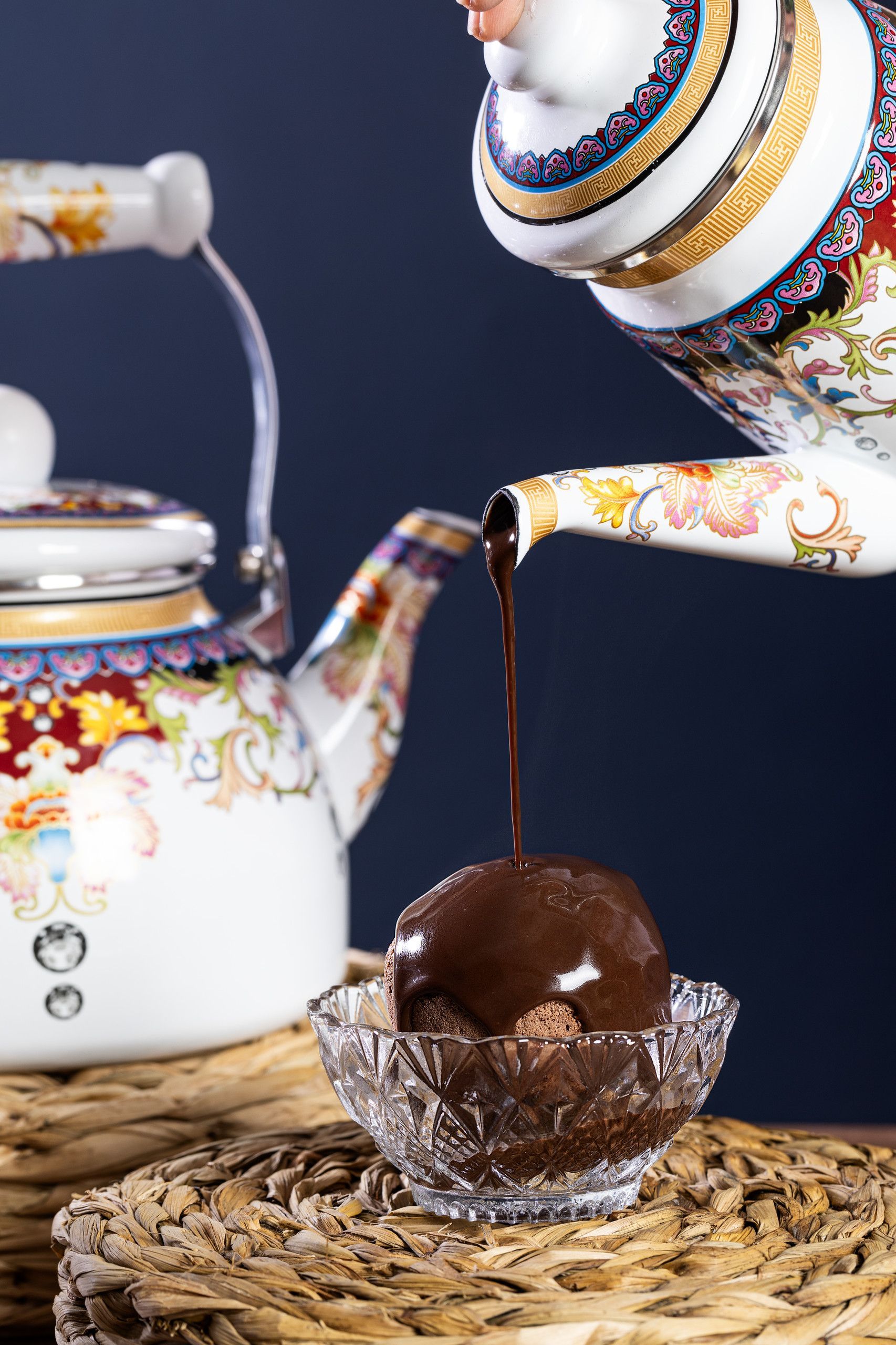 Bule servindo chá em uma xícara sobre uma esteira trançada, com uma trufa de chocolate à frente.