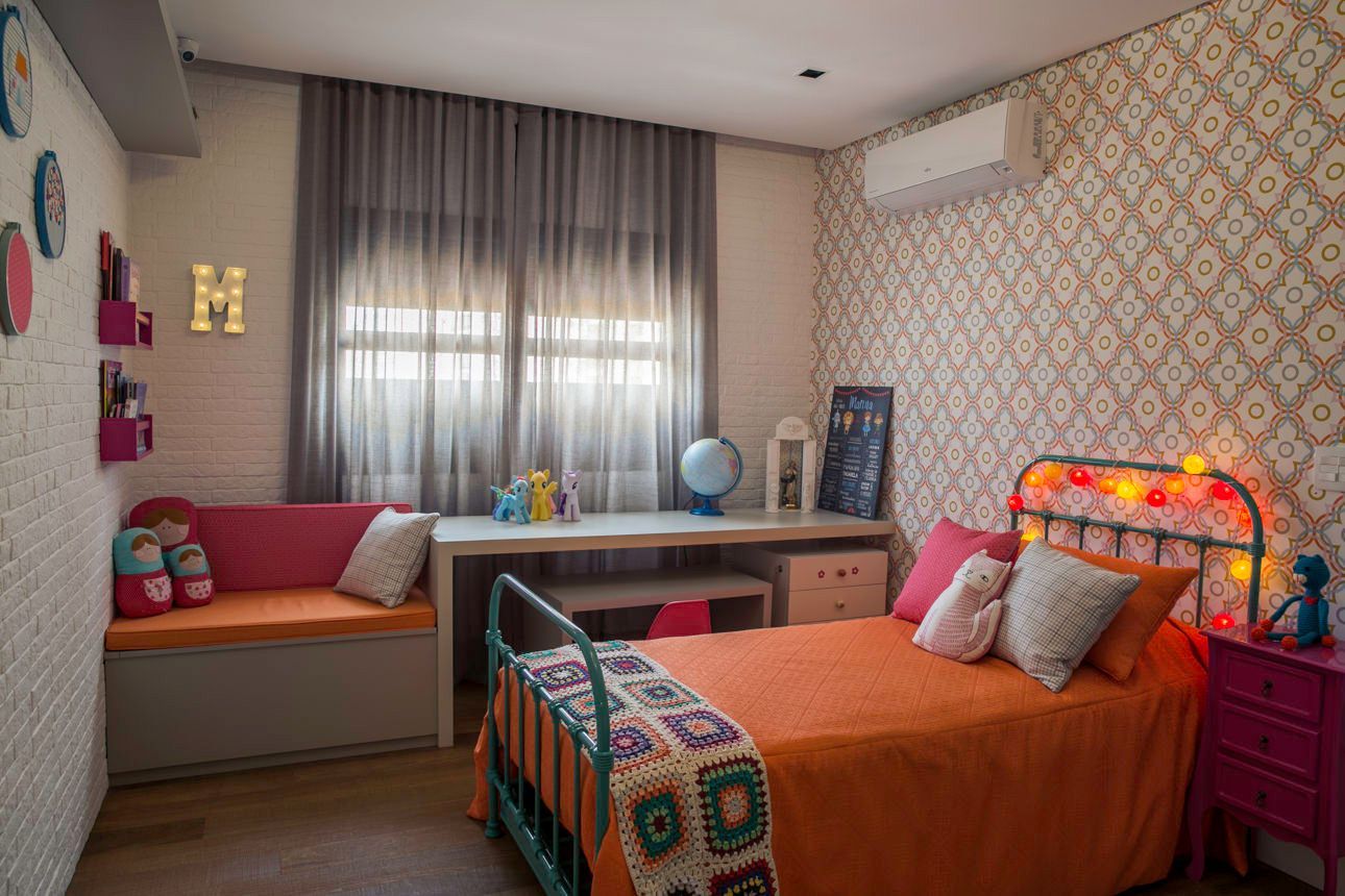 Quarto aconchegante com colcha laranja, banco junto à janela, escrivaninha e papel de parede estampado.