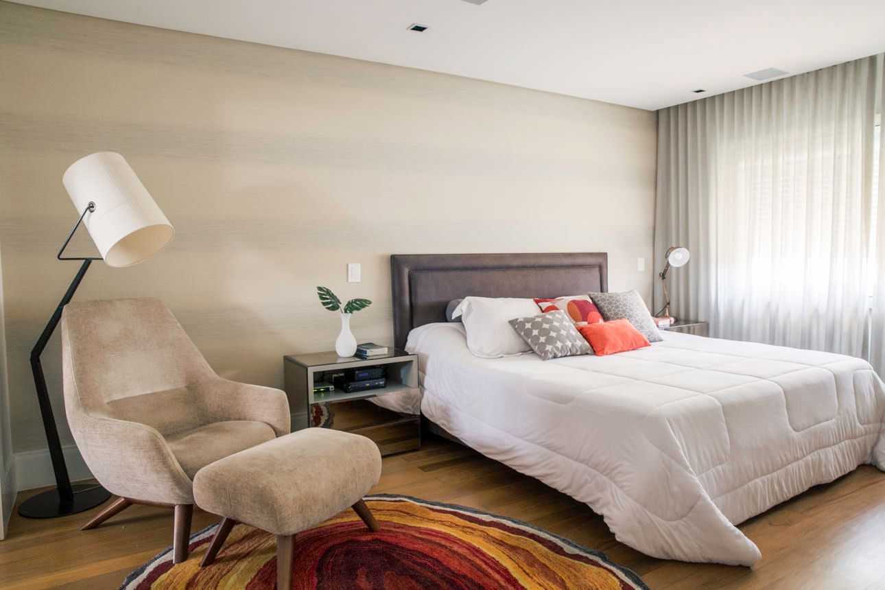 Quarto luminoso com uma cama grande, poltrona bege, luminária de chão e cortinas translúcidas junto à janela.