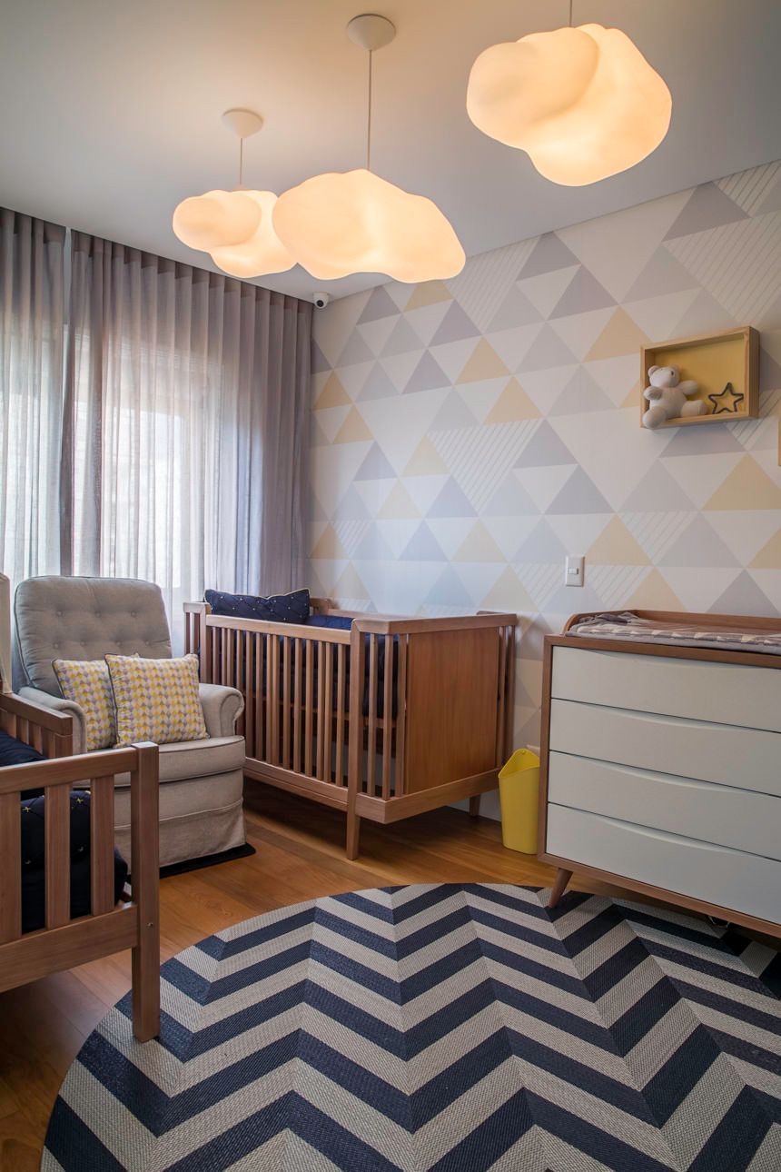 Quarto de bebê aconchegante com dois berços, tapete estampado, luminárias de teto em formato de nuvem e papel de parede geométrico.