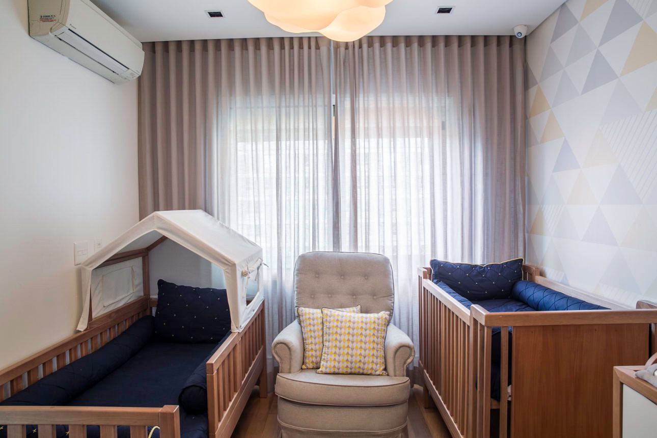 Quarto de bebê com dois berços, uma poltrona almofadada e cortinas translúcidas junto a uma janela bem iluminada.
