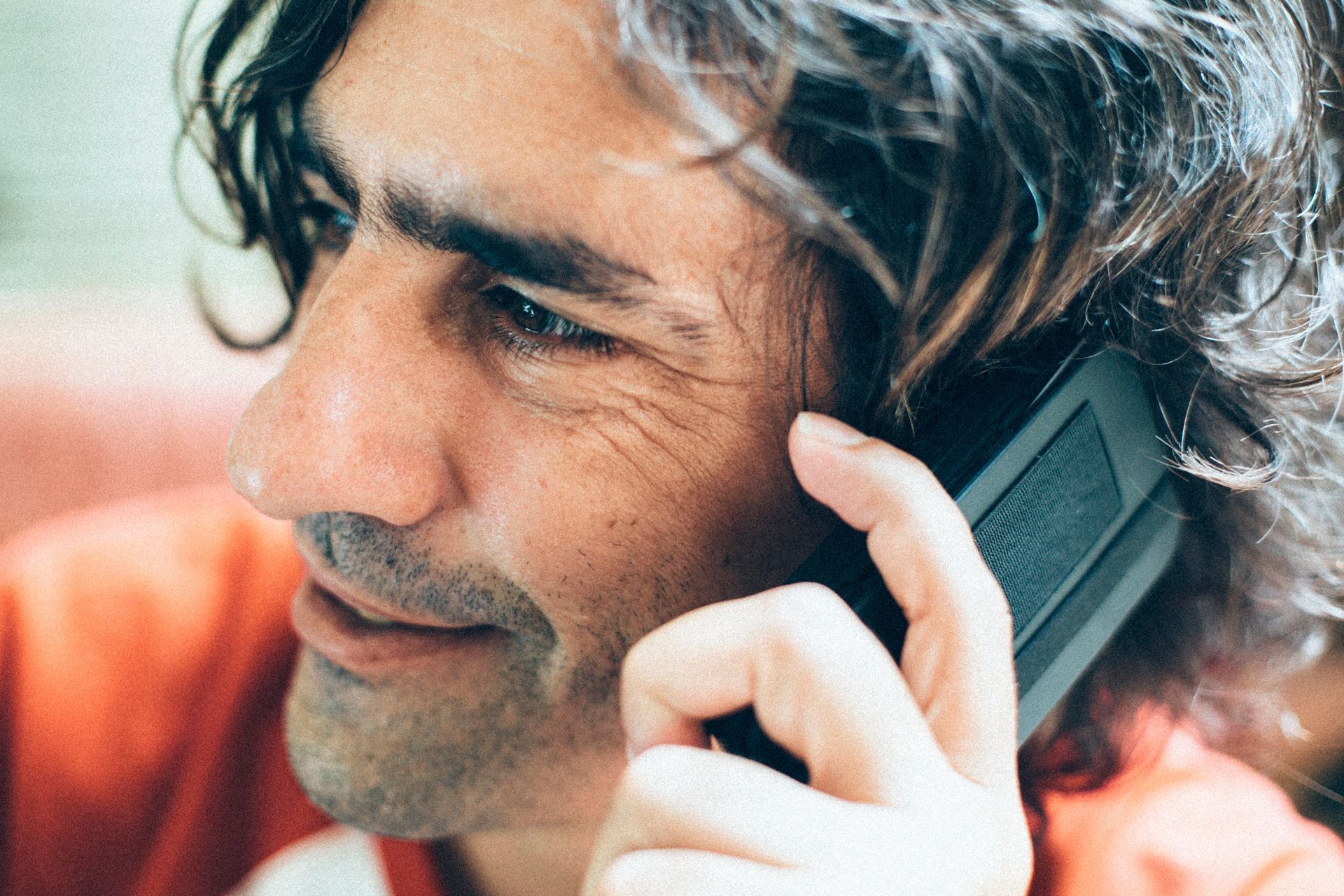 Pessoa sorridente de cabelos grisalhos segurando um telefone junto à orelha em ambiente interno.
