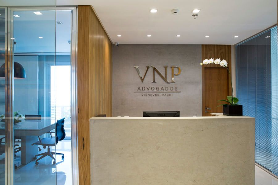 Recepção moderna de escritório de advocacia com balcão bege, painéis de madeira e paredes de vidro, com a marca “VND Advogados”.