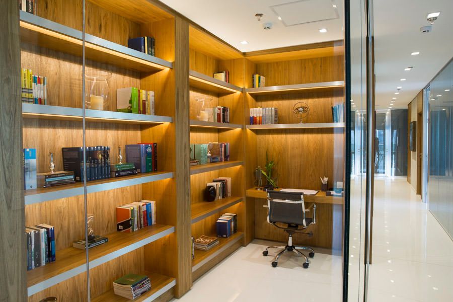 Biblioteca de escritório moderna com estantes de madeira, livros e uma mesa em um corredor com paredes de vidro.