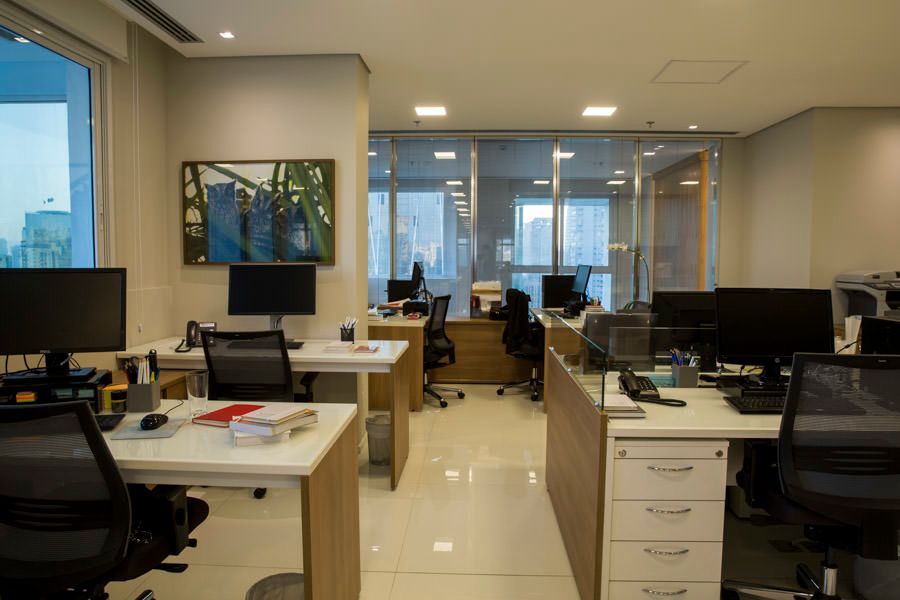 Interior de escritório moderno com mesas, computadores, cadeiras e grandes janelas ao fundo.