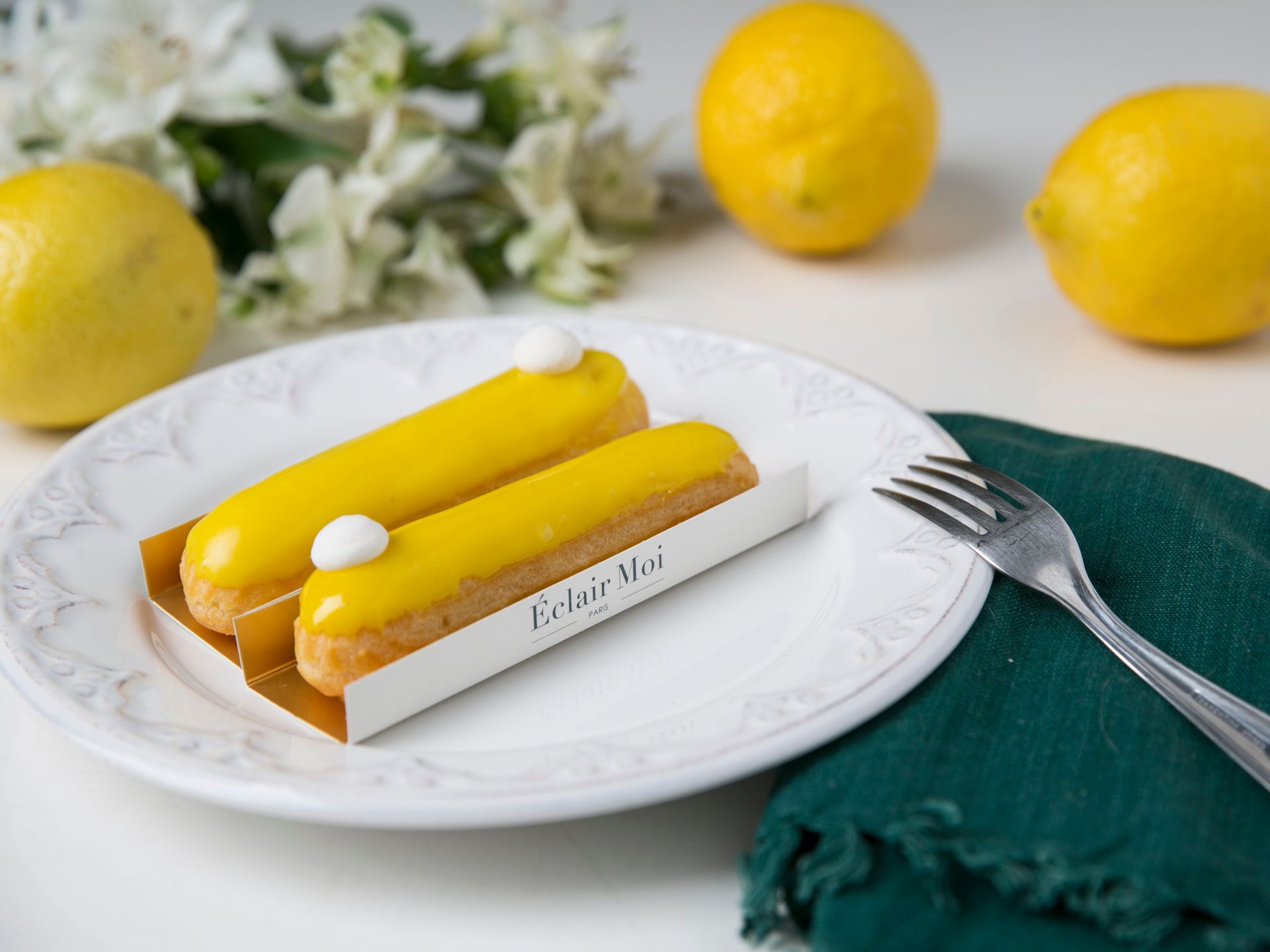 Tarte de limão num prato branco com limões inteiros, flores e um garfo sobre um guardanapo verde.