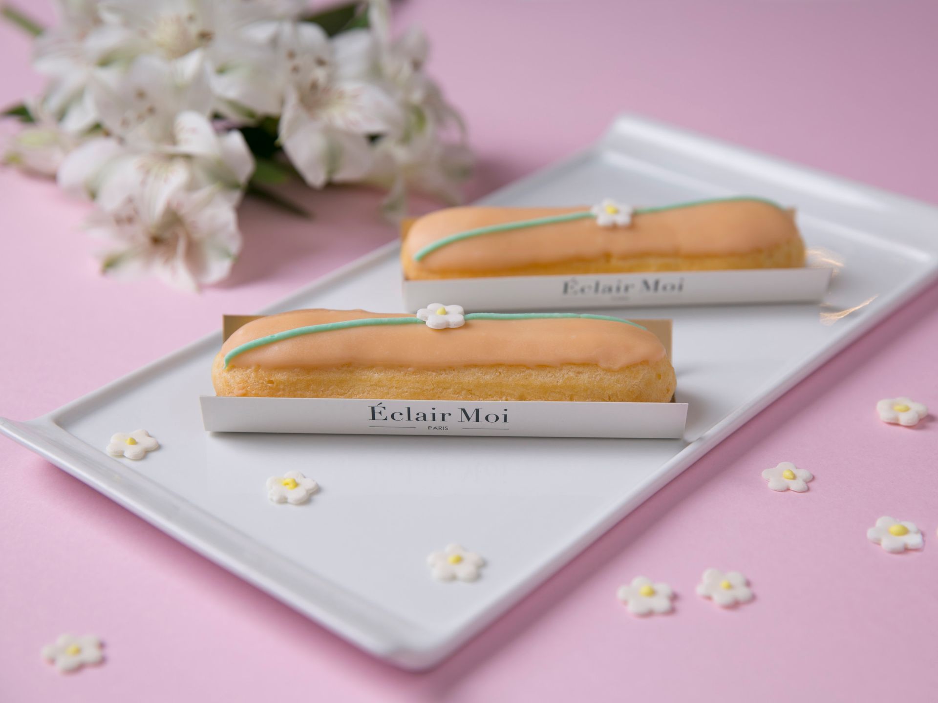 Duas éclairs em tons pastel sobre uma bandeja branca com flores, em um fundo rosa.