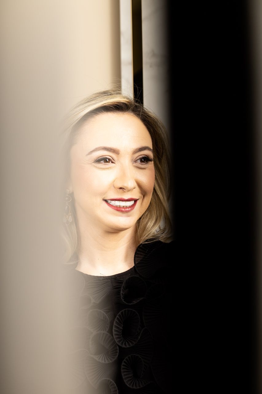 Mulher sorridente de cabelos loiros e blusa preta, em pé ao lado de uma cortina clara.