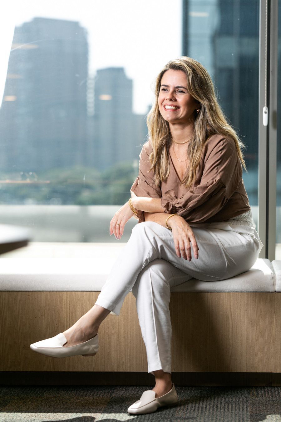 Mulher sentada no parapeito de uma janela, vestindo roupas claras e sorrindo, com prédios da cidade desfocados ao fundo.