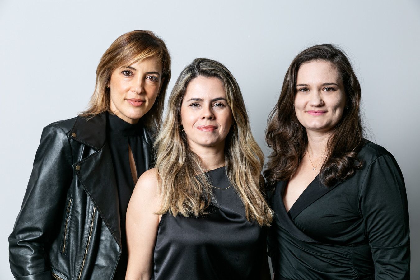 Três mulheres posando juntas com roupas pretas contra um fundo cinza claro.