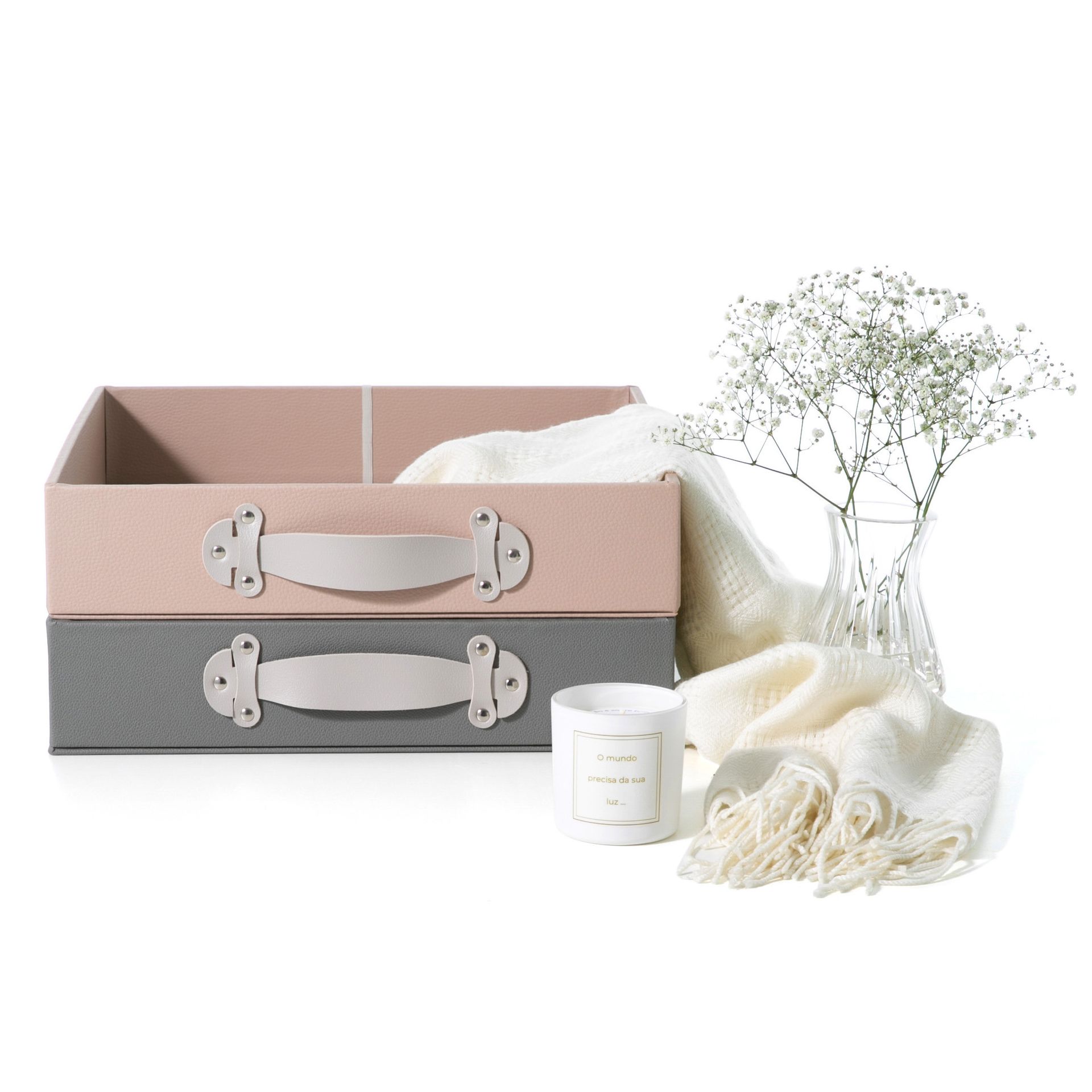 Duas caixas organizadoras em tons pastel com alças ao lado de uma manta branca, uma vela e flores secas sobre um fundo branco.