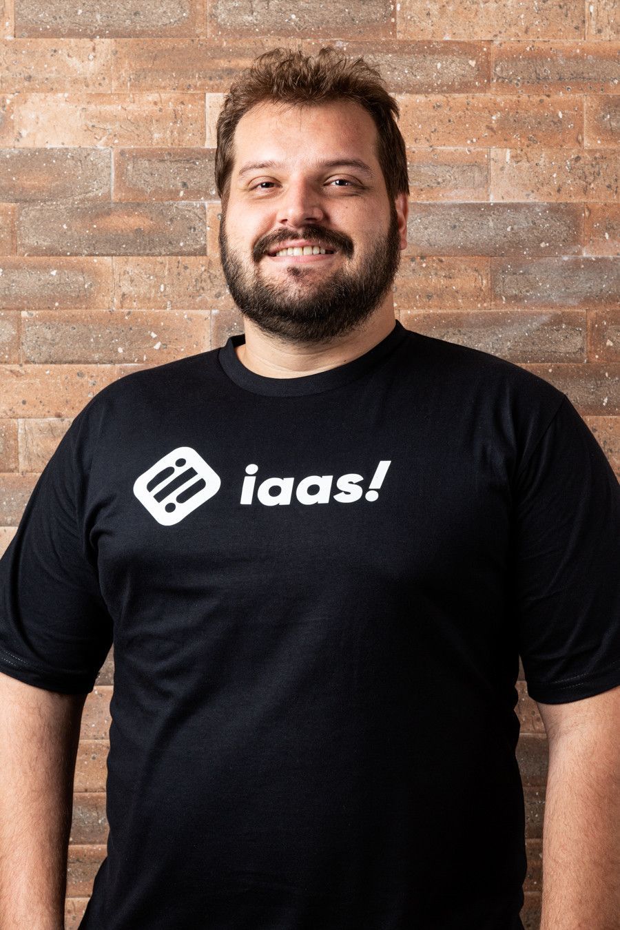 Homem sorridente vestindo uma camiseta preta da iaas! está encostado em uma parede de tijolos.
