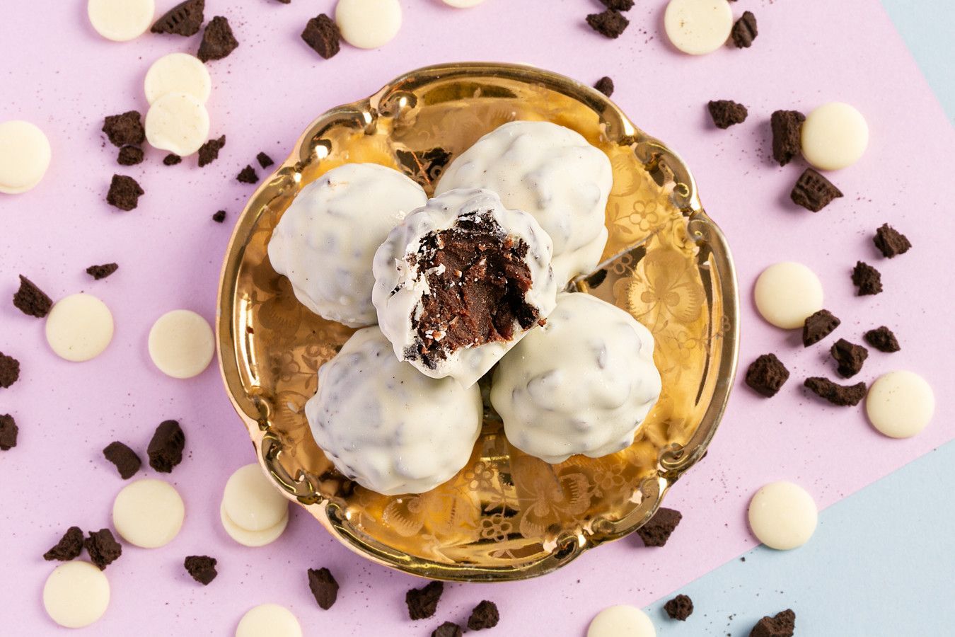 Biscoitos de merengue branco em uma tigela dourada sobre um fundo rosa com gotas e migalhas de chocolate espalhadas.