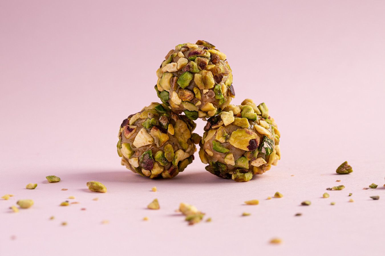 Três trufas de chocolate cobertas com pistache, empilhadas sobre um fundo rosa, com migalhas de nozes espalhadas.