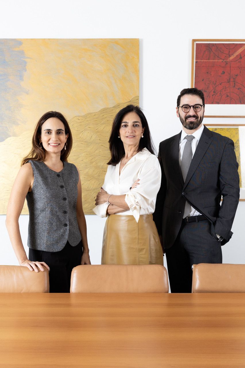Três profissionais em pé atrás de uma mesa de conferência em um escritório com obras de arte emolduradas na parede.