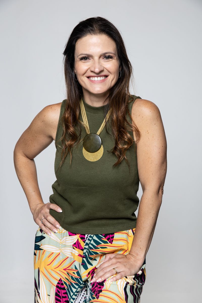 Mulher sorridente, vestindo uma blusa verde-oliva sem mangas e uma saia colorida, em pé com uma das mãos no quadril.