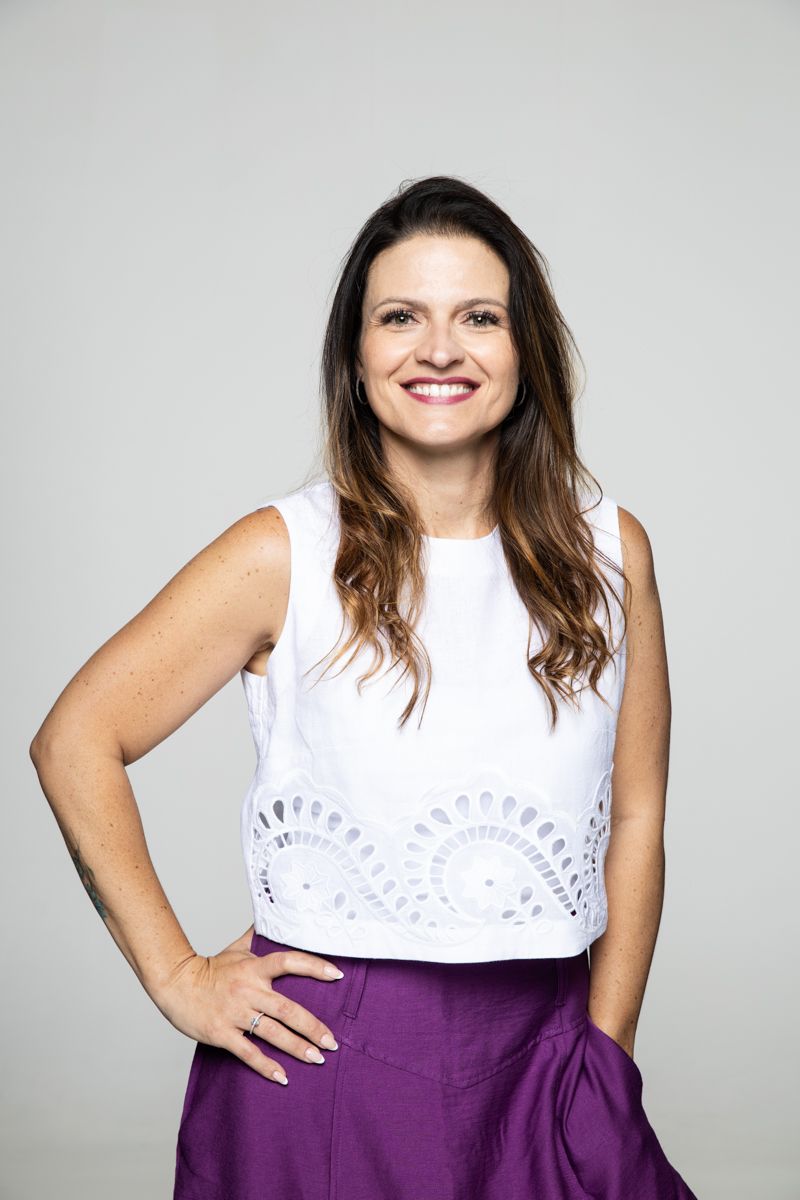 Mulher com blusa branca sem mangas e calça roxa, sorrindo com a mão na cintura contra um fundo liso.