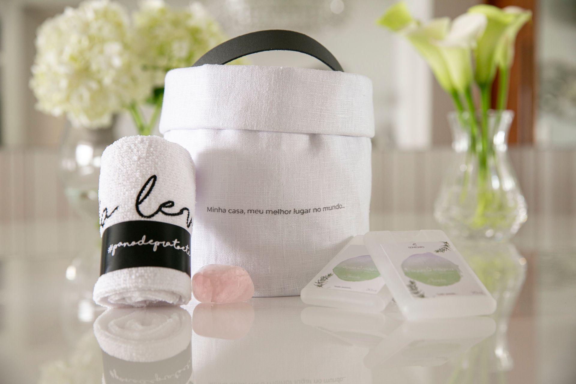 Kit de spa com cesta de toalhas brancas, vela, sabonetes e flores sobre uma mesa iluminada.