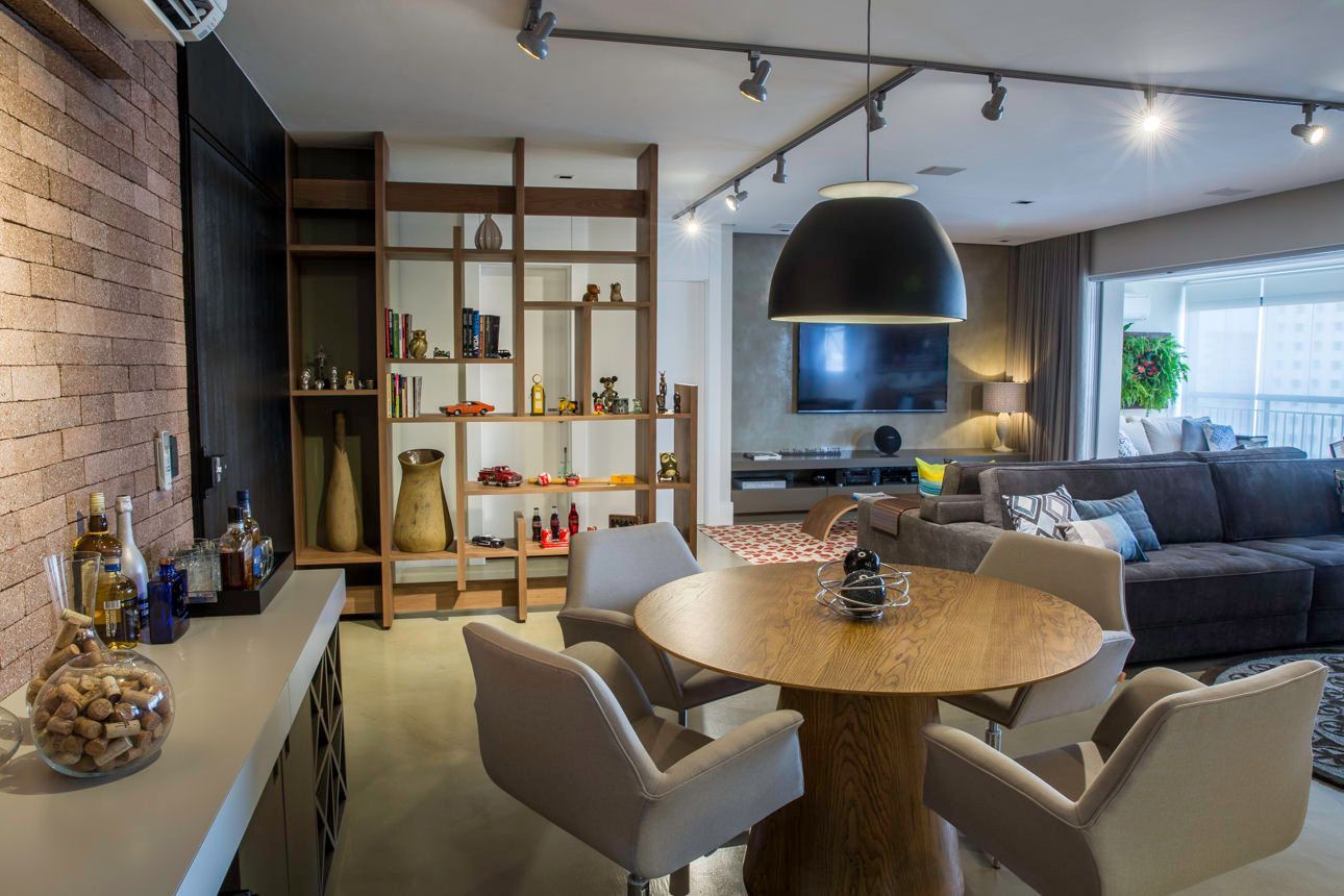Sala de estar moderna com mesa de jantar redonda, cadeiras bege, sofá escuro, parede de tijolos à vista e janela ampla.