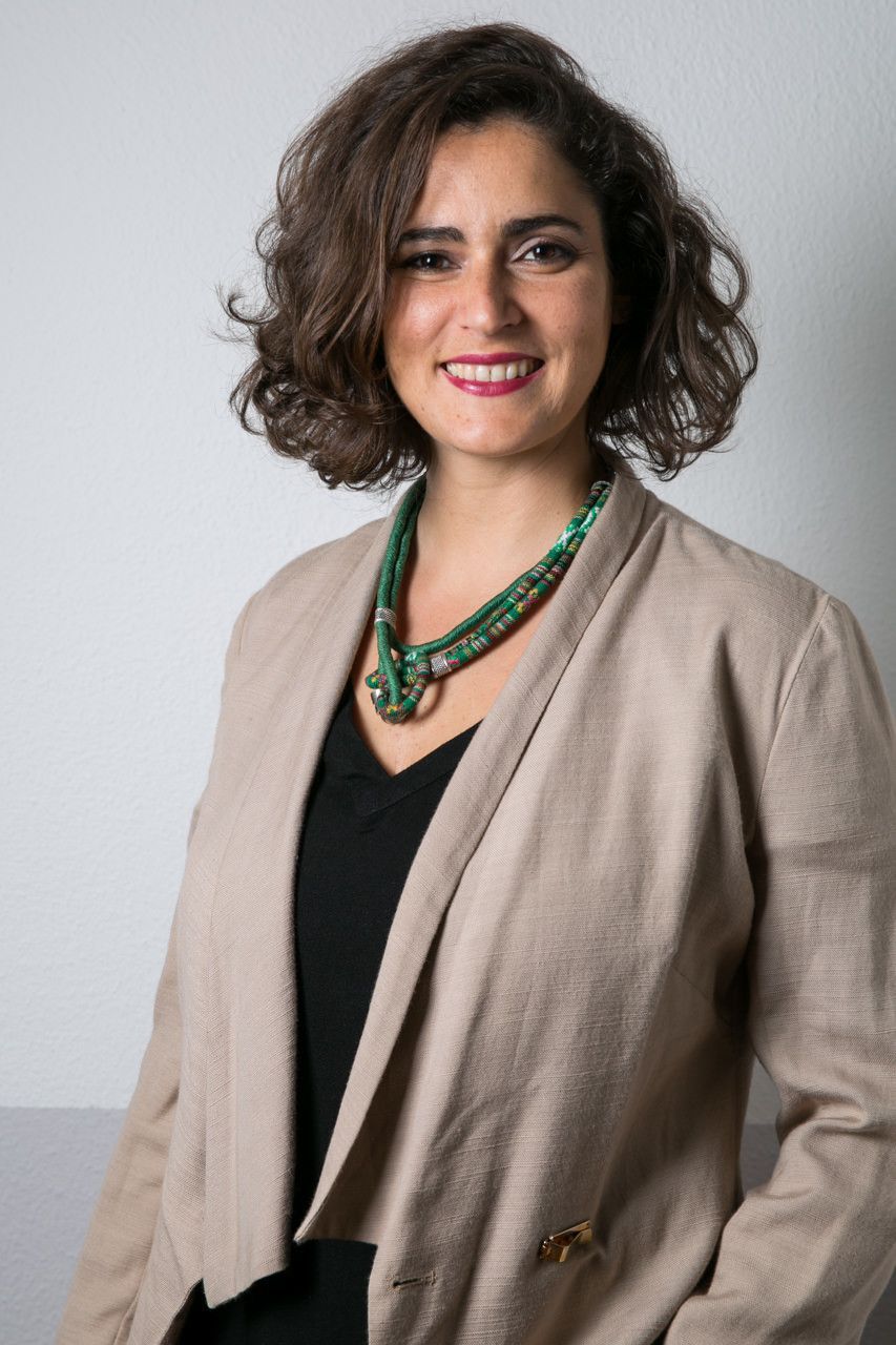 Mulher com blazer bege e colar verde, sorrindo contra um fundo cinza claro.