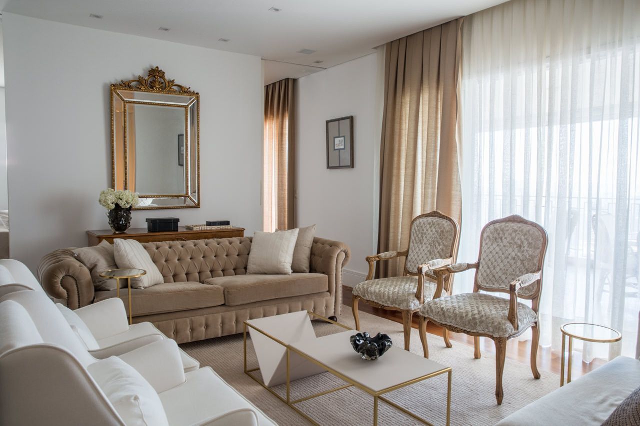 Sala de estar elegante com sofás bege, espelho dourado e cortinas translúcidas junto a uma janela bem iluminada.
