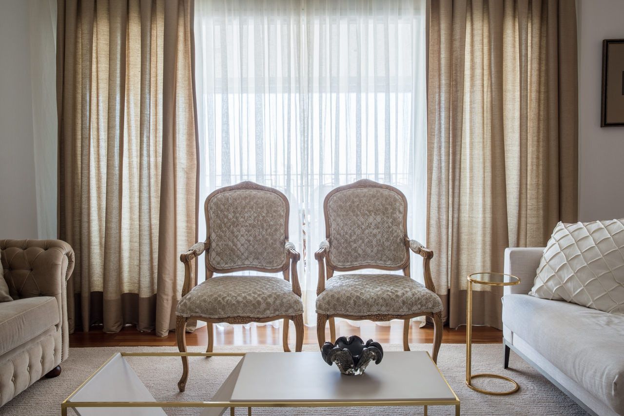 Sala de estar elegante com duas cadeiras ornamentadas, cortinas amplas nas janelas e uma mesa de centro decorada.