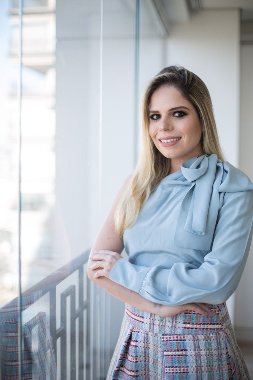 Mulher com blusa azul-clara, sorrindo, em pé junto a um corrimão em um corredor interno bem iluminado.