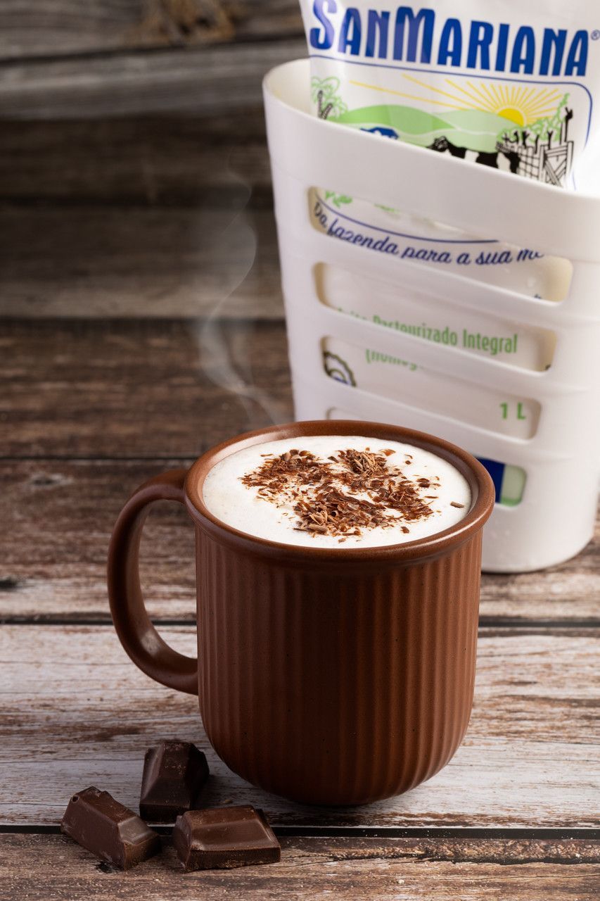 Caneca marrom de cappuccino com raspas de chocolate ao lado de quadradinhos de chocolate e um porta-pacote branco.