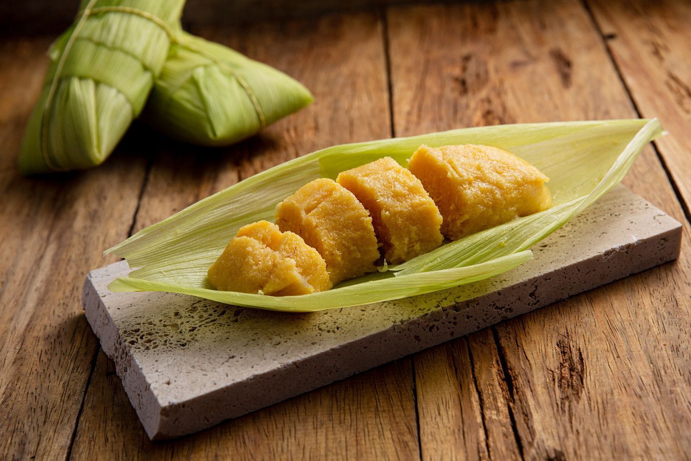 Bolinhos de arroz dourados em uma travessa de pedra forrada com folhas verdes, envoltos em folhas dobradas, sobre uma mesa de madeira.