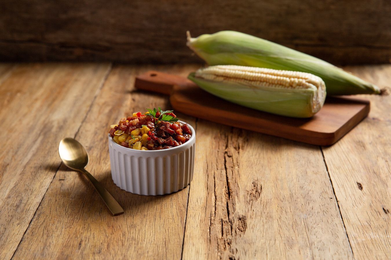 Milho grelhado em uma tábua de madeira ao lado de uma tigela de chili e uma colher em uma mesa rústica.