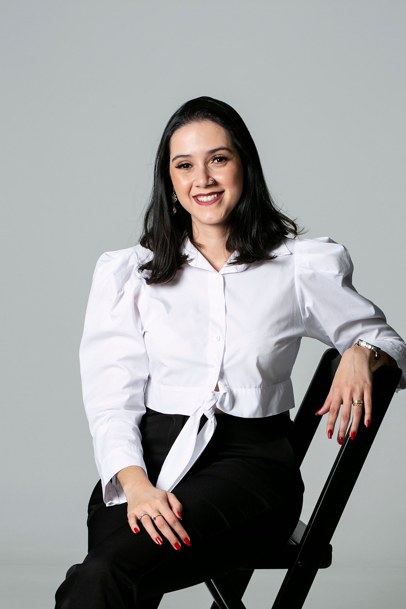 Mulher de blusa branca e calça preta sentada em uma cadeira contra um fundo cinza, sorrindo para a câmera.