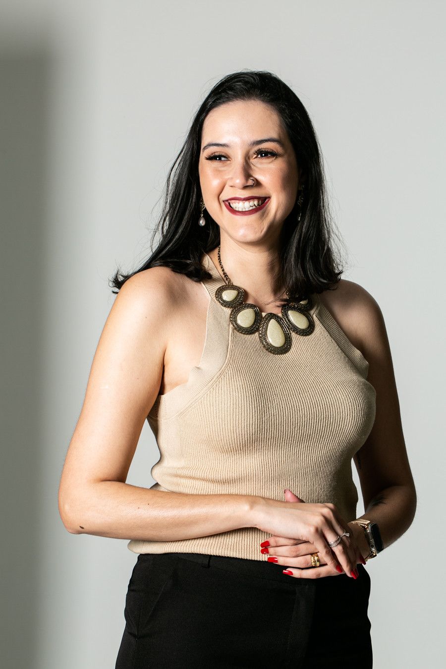 Mulher sorridente vestindo um top bege de alças e calças pretas, em pé contra um fundo cinza liso.
