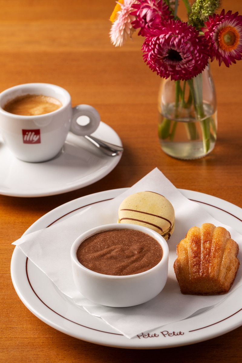 Café e doces num prato branco ao lado de uma xícara e um vaso de flores sobre uma mesa de madeira.