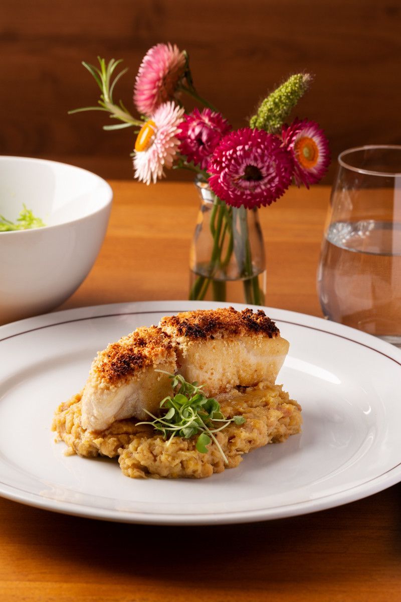 Bolinho de peixe grelhado com verduras em um prato branco, ao lado de um copo d'água e flores sobre uma mesa de madeira.