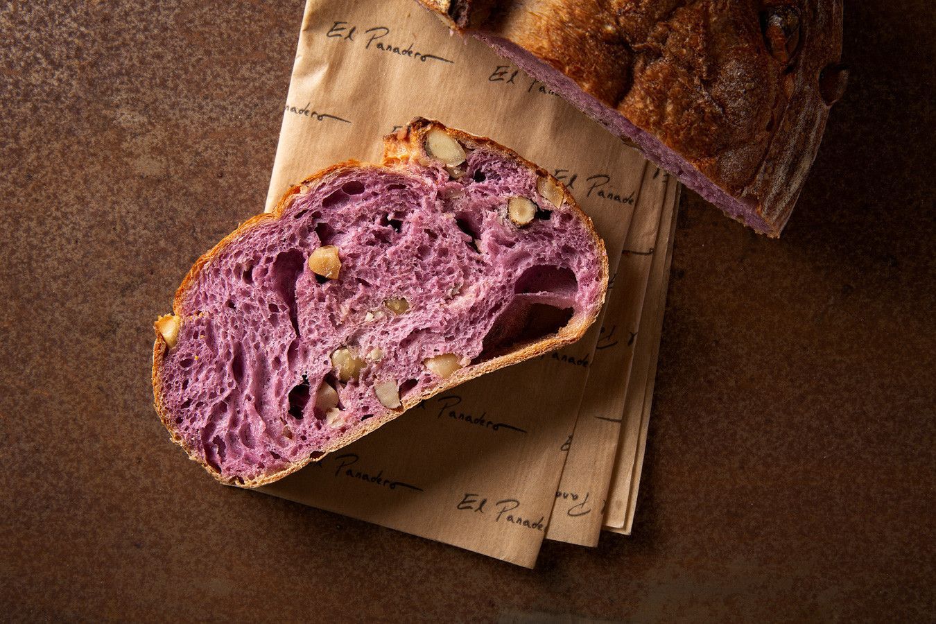 Pão rústico roxo com nozes sobre papel pardo, fatiado para mostrar o miolo.