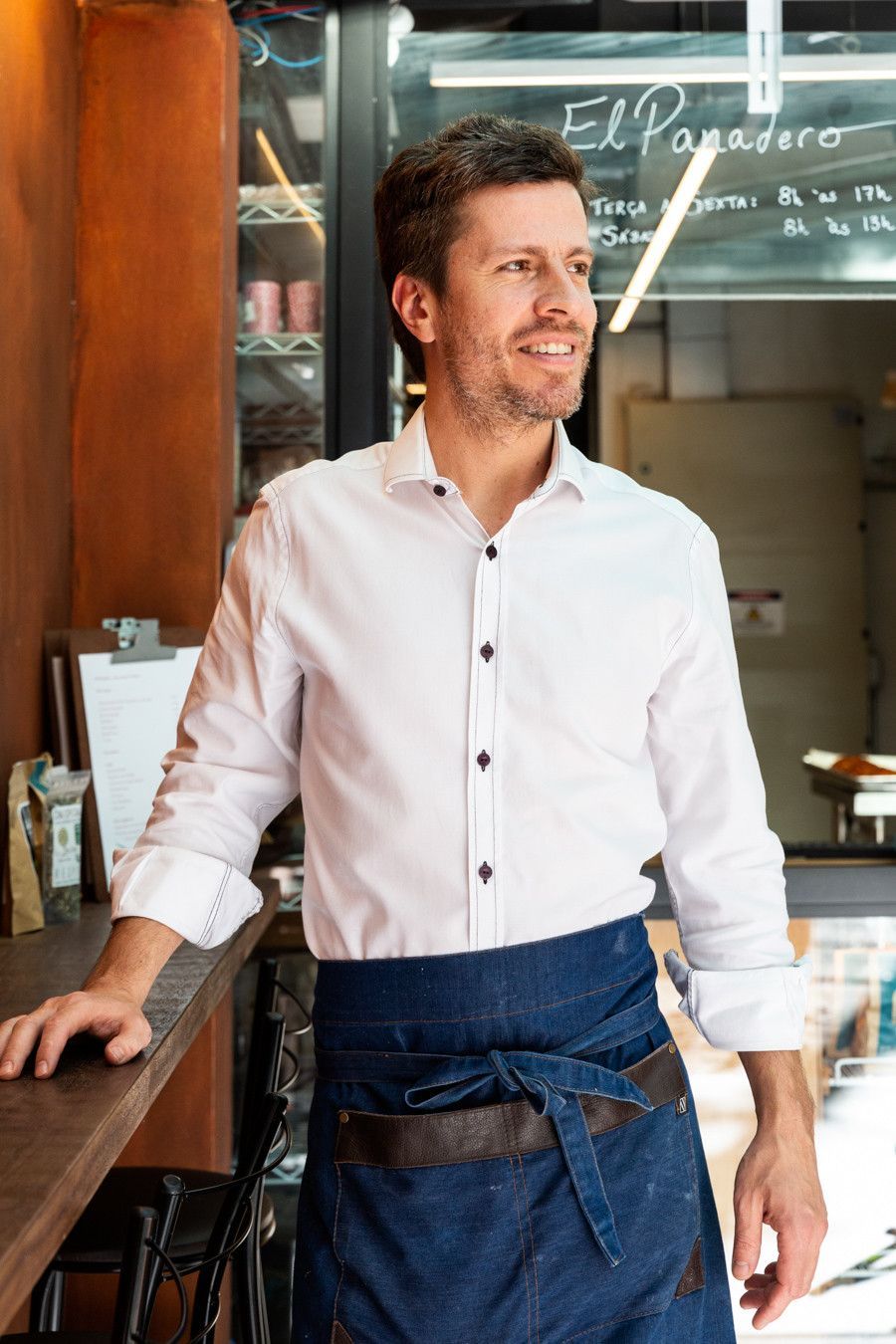 Homem de camisa branca e avental azul parado na porta de um café, sorrindo e olhando para o lado.