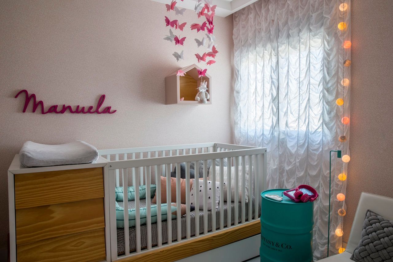 Quarto de bebê com berço branco, cômoda de madeira, decoração de parede com borboletas rosa e cortinas leves iluminadas por luzes de cordão.
