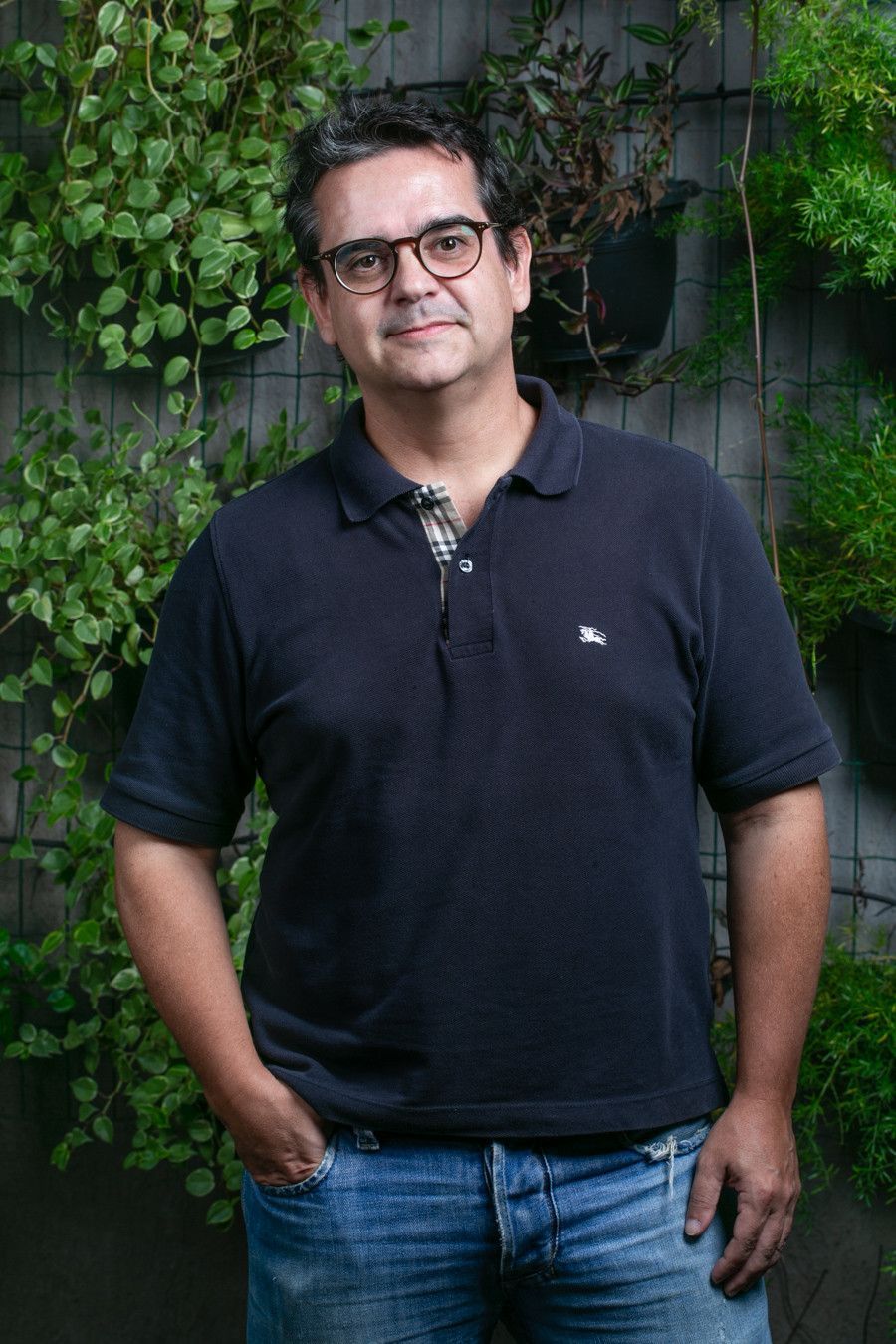 Homem de pé ao ar livre, vestindo uma camisa polo preta e calça jeans, encostado em uma parede coberta de trepadeiras.