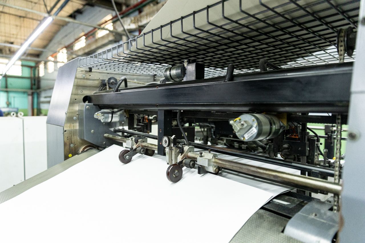Grande impressora industrial alimentando uma folha branca através de uma máquina de metal em uma fábrica.
